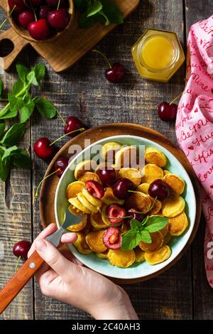 In den Händen einer Frau Frühstück. Mini-Pfannkuchen mit Kirschen und Honig auf einem rustikalen Holztisch. Draufsicht flach lagen Hintergrund. Stockfoto