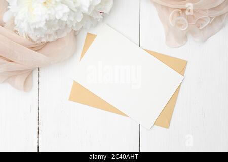 Romantisches Liebesbrief Konzept. Flacher Kraftpapier-Umschlag, leere Notiz, Pfingstrosen-Blumen und beiges Tuch auf weißem Holztisch. Draufsicht mit Kopierbereich Stockfoto