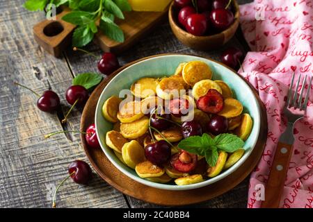 Winziges Pfannkuchen-Frühstück. Mini-Pfannkuchen mit Kirschen und Honig auf rustikalem Holztisch. Speicherplatz kopieren. Stockfoto