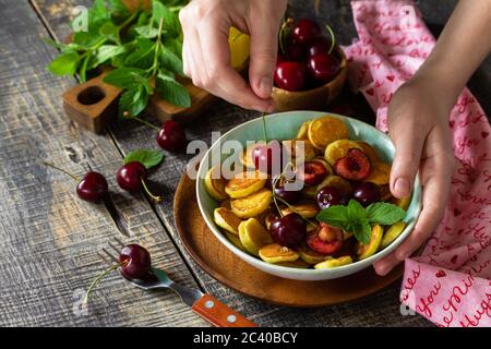 In den Händen einer Frau Frühstück. Mini-Pfannkuchen mit Kirschen und Honig auf einem rustikalen Holztisch. Stockfoto