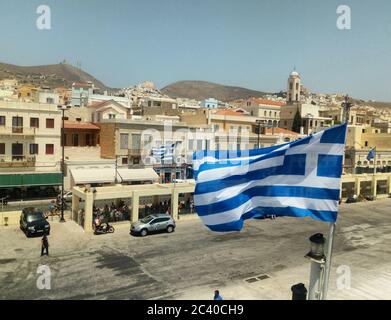 Griechische Flagge flattert in der Meeresbrise vom Fährschiff auf der schönen, traditionellen griechischen Insel Hydra.der charmante Hafen im Hintergrund Stockfoto