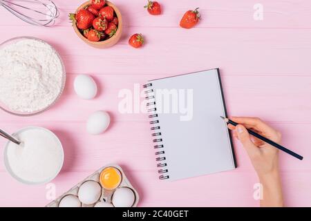 Hausgemachte Erdbeerkuchen Rezept, Frau Hand schreiben in leerem Notizbuch neben den Zutaten frische Beeren, Eier, Mehl und Zucker auf rosa Tisch, Fla Stockfoto