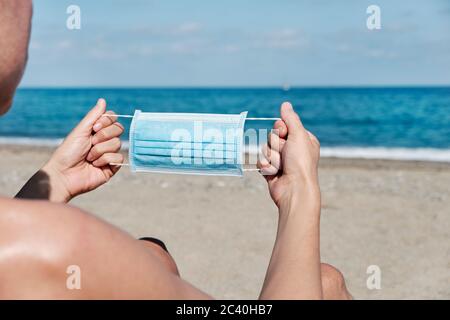 Nahaufnahme eines jungen Kaukasiermannes, der in einem blauen Liegestuhl am Strand sitzt, eine OP-Maske an- oder auszieht, mit dem Meer im Hintergrund Stockfoto