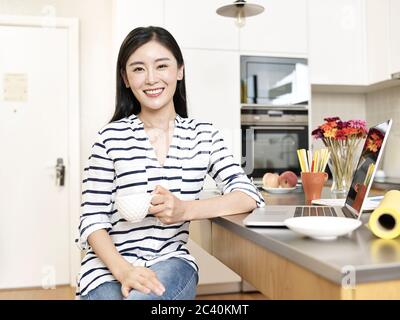 Portrait eines glücklichen jungen asiatischen freiberuflichen Designer von zu Hause aus sitzen an der Küchentheke mit einer Tasse Kaffee Blick auf Kamera lächelnd Stockfoto