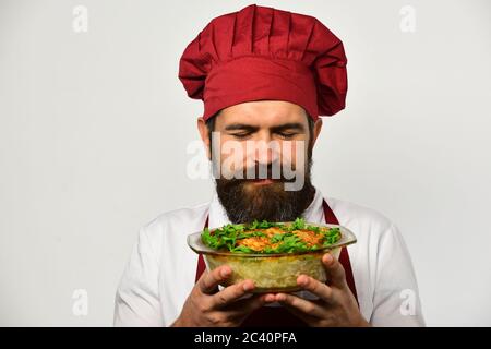 Kochen und vegetarische Ernährung Konzept. Der Koch hält eine Schüssel mit Kartoffelauflauf und Petersilie. Kochen Sie mit zufriedenen Gesicht in burgunderrot Uniform riecht gebackene Gericht. Mann mit Bart isoliert auf weißem Hintergrund Stockfoto