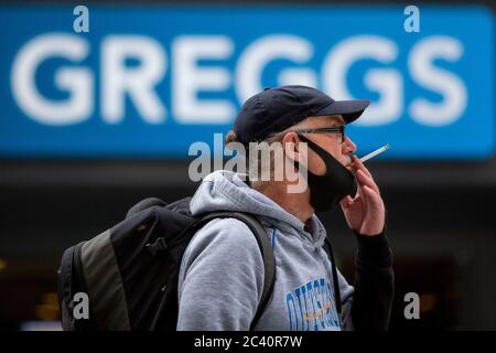 Ein Mann raucht eine Zigarette, während er eine Gesichtsmaske über seinem Kinn vor einem Greggs-Laden in Cardiff trägt, 22. Juni 2020. Stockfoto