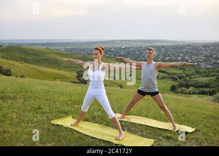 Ein Mann und eine Frau praktizieren synchron Yoga, meditieren Gleichgewicht in der Pose des stehenden Grases auf der Natur. Stockfoto