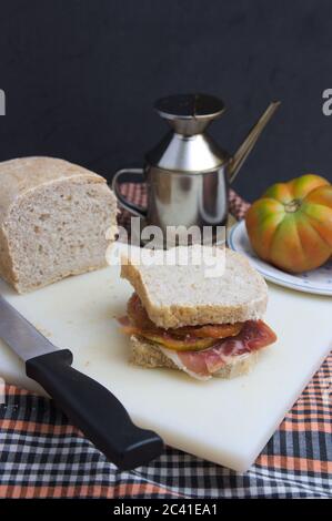 Stillleben eines serrano Schinken Sandwich mit Tomaten und Olivenöl, im Hintergrund sehen wir die Zutaten und ein Messer zum Schneiden von Brot. Dunkler Hintergrund Stockfoto