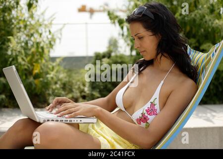Junge dunkelhaarige Frau im Bikini auf einem Liegestuhl tippt auf einem Laptop - Flexibilität - Sommer Stockfoto
