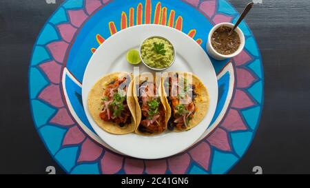 Draufsicht auf einen weißen Teller mit drei mexikanischen Tacos Fleisch, begleitet von einem kleinen Topf Guacamole und würziger Sauce auf einem Holztisch mit verschiedenen Colos Stockfoto