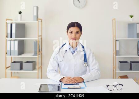 hispanic Ärztin beim Betrachen der Kamera Hört einem Patienten zu, benutzt eine Webcam-Konferenz, während er an einem Tisch im Büro einer medizinischen Klinik sitzt. Stockfoto
