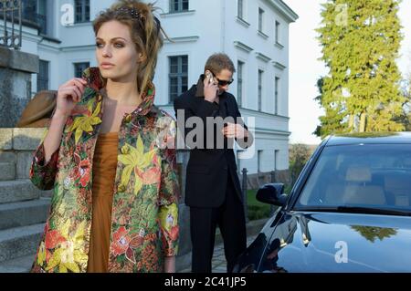 Junge manipulierte Frau steht vor einem elegant gekleideten Mann, der am Telefon ist - Anwesen - Reichtum - Leibwächter Stockfoto