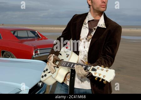 Der junge Mann steht hinter zwei Oldtimern und spielt Gitarre - Strand - Meer Stockfoto