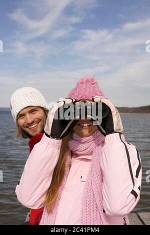 Junge Frau in Winterkleidung neben einem Mann schaut durch Fernglas - Freizeit - See Stockfoto