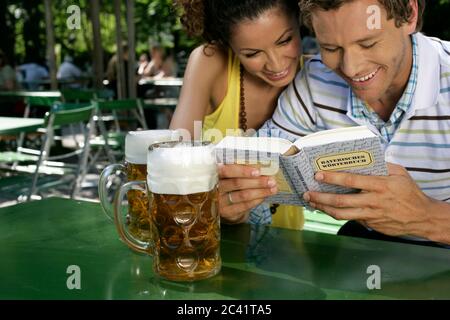Junge Frau mit lockigen Haaren sitzt neben einem Mann und schaut in ein Buch - Sommer Flirt - Biergarten Stockfoto