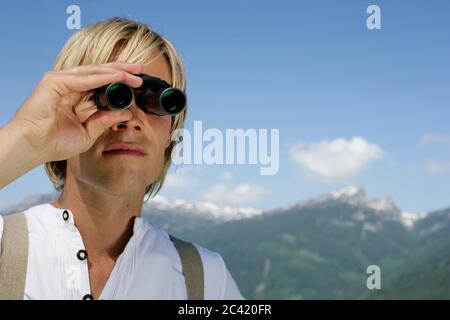 Der junge, blonde Mann blickt mit einem Fernglas in die Ferne in eine Berglandschaft Stockfoto