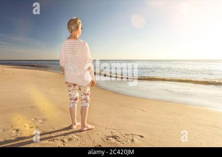 Ältere Frau, die am Strand steht und mit einer malerischen Linse auf das Meer blickt Stockfoto