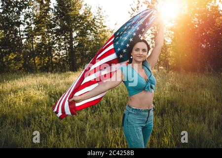 Frau trägt Jeans und blaues Top hält eine amerikanische Flagge über ihrem Kopf auf einer Natur während der Sonnenuntergang Feier. Stockfoto