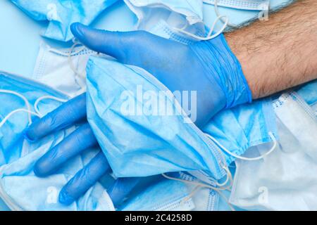 Mans Hand in Gummi blau Handschuh hält verwendet Einweg-OP-Maske auf einem Hintergrund von anderen Gesichtsmasken. Medizinischer Müll nach einer Coronavirus-Pandemie Stockfoto