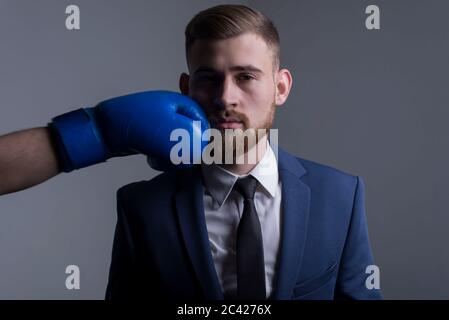 Nahaufnahme Porträt eines jungen bärtigen Kerl in einem Business-Anzug, Geschäftsmann, Seitenansicht die Hand eines Gegners in einem Boxhandschuh schlägt. Der Moment von Stockfoto