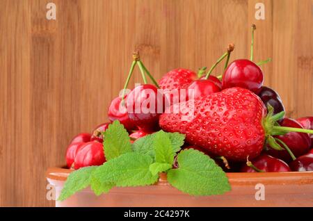 Frühlingsfrüchte, frische Kirschen, Erdbeeren mit Zitronenmelisse in rustikaler, Keramikschale vor Holzgrund, Seitenansicht Stockfoto