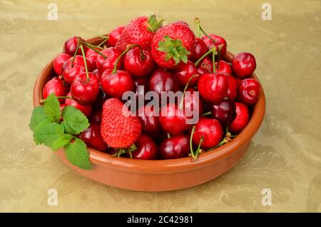 Reife, rote Erdbeeren und Kirschen in rustikaler Keramikschale auf einem mit goldenem Öltuch bedeckten Tisch, verziert mit frischen Zitronenmelisse-Blättern Stockfoto