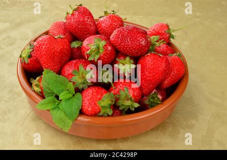 Reife, rote Erdbeeren in rustikaler Keramikschale auf einem mit goldenem Öltuch bedeckten Tisch, verziert mit frischen Zitronenmelisse-Blättern Stockfoto