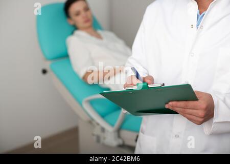 Patientin sitzt auf dem Behandlungstisch, der Arzt im Vordergrund notiert sich Stockfoto