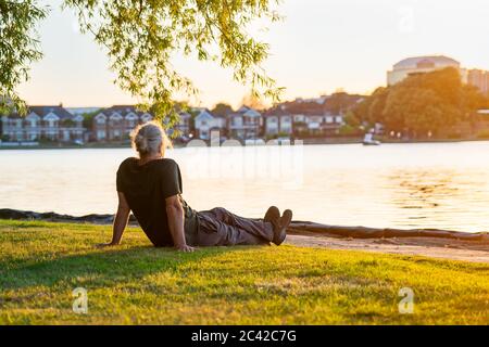 Rückansicht graues Haar reifer Mann sitzt auf dem grünen Gras am Ufer des Parks See und genießen den Sonnenuntergang. Ein einfaches Vergnügen für die geistige Gesundheit. Nat Stockfoto