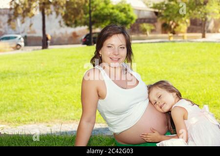 Nettes kleines Mädchen umarmt und küsst und hört ihre Mütter schwangeren Bauch sitzen im Freien auf einer grünen Wiese in einem Stadtpark auf einer sonnigen da Stockfoto