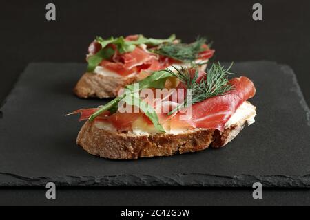 Buchweizen-Toasts mit Ziegenkäse, Prosciutto und Rucola liegen auf einem schwarzen Steinbrett auf einem schwarzen Tisch. Nahaufnahme Stockfoto