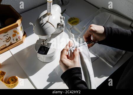 High-Winkel Nahaufnahme von unkenntlich weiblichen Nähen Nylon elastischen Band zu Stoff mit Nadel und Fäden, während die Schaffung von Schutz Gesichtsmaske zu Hause Stockfoto