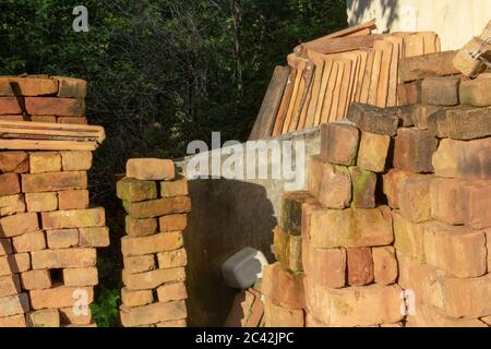 Haufen von Ziegelsteinen, die in der Nähe eines alten Hauses mit einem weiteren Haufen von Ziegelsteinen und Bäumen auf dem Hintergrund ruhen Stockfoto