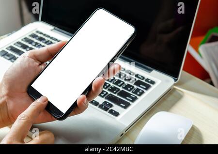 Man Hand drückt das Smartphone mit einem leeren Bildschirm für die Gestaltung oder Werbung auf dem Schreibtisch und den Laptop-Computer auf dem Arbeitstisch. Stockfoto