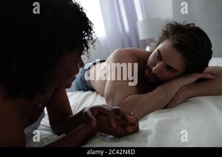 Entspannter junger Mann, der auf dem Bett liegt und der afroamerikanischen Freundin zuhört, während er sich zu Hause auf dem Bett abkühlt Stockfoto