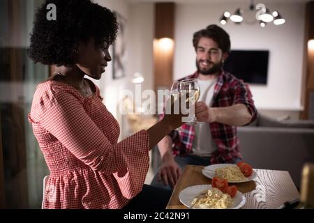 Seitenansicht der glücklichen schwarzen Frau lächelnd und klirrende Weingläser mit Freund, während mit romantischen Abendessen in gemütlichen Wohnung zusammen Stockfoto