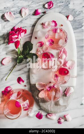 Erfrischendes kaltes Getränk im Sommer. Flache Rosenlimonade mit Eis in Gläsern und Krug und frischen Rosenblütenblättern auf dem Brett über grauem Marmor ta Stockfoto