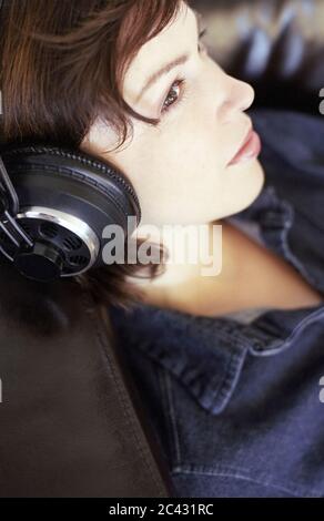Eine junge dunkelhaarige Frau sitzt bequem auf einem Ledersessel und hört Musik mit Kopfhörern - Freizeit - Unterhaltung Stockfoto