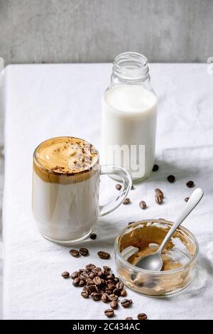 Dalgona schaumig Kaffee Trend koreanisch trinken Milch Latte mit Kaffee-Schaum in Glas Becher, dekoriert mit gemahlenem Kaffee auf weißem Baumwolltischtuch. Zutaten Stockfoto