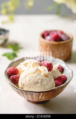 Hausgemachtes Karamell-Vanille-Eis mit gefrorenen Himbeeren in Keramikschale auf weißem Marmortisch mit Blumen dahinter. Stockfoto