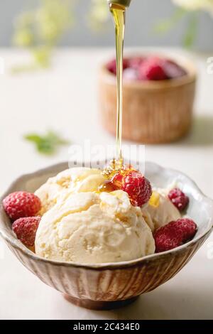 Hausgemachtes Karamell-Vanille-Eis mit fließendem Sirup, gefrorene Himbeeren in Keramikschale auf weißem Marmortisch mit Blumen dahinter stehend. Stockfoto