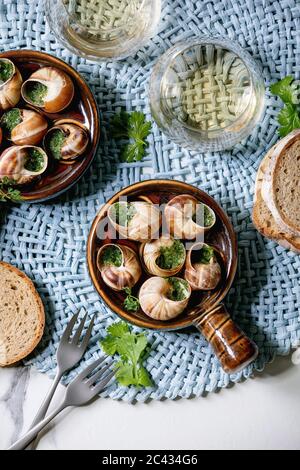 Escargots de Bourgogne - Schnecken mit Kräuterbutter, Gourmet-Gericht, in traditioneller Keramikpfanne mit Koriander, Brot, Glas Weißwein auf blauer Serviette Stockfoto