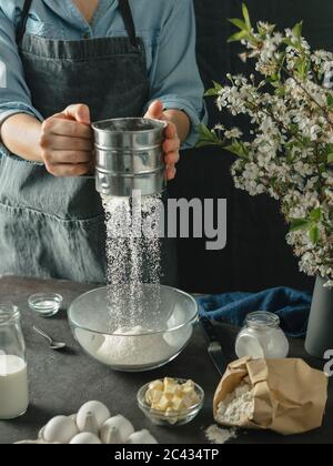 Junge Frau, die Kuchen macht. Weibliche Hände sieben Mehl aus Metallsieb in Glas Rührschüssel auf dunklem Hintergrund. Lebensmittelzutaten und Küchengeschirr mit Stockfoto