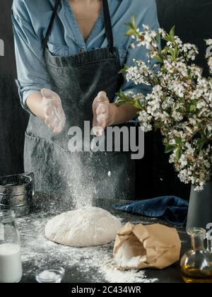 Junge Frau in blauem Hemd und grauer Schürze, die Brotteig macht. Weibliche Hände Kneten Teig in der dunklen Küche. Schwarzer Tisch mit Zutaten für Lebensmittel - Mehl, m Stockfoto