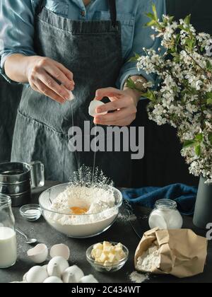 Junge Frau in blauem Hemd und grauer Schürze, die Kuchen macht. Weibliche Hände brechen das Ei in Mischen boul mit Mehl auf dunklem Hintergrund. Zutaten für Lebensmittel und KI Stockfoto