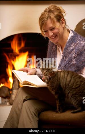 Frau mit rotblonden Haaren sitzt auf einem Sessel vor dem Kamin mit einem Buch und streichelt die Katze - Freizeit - entspannend Stockfoto