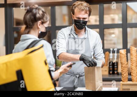 Weibliche Kurierin nimmt Online-Bestellung für die Lieferung an der Theke mit Verkäufer in der kleinen Konditorei oder Café. Konzept der Mitnehmen von Lebensmitteln während einer Pandemie Stockfoto