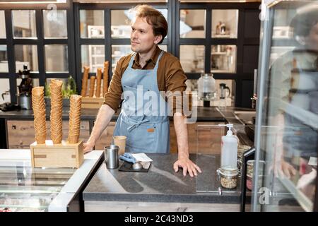Porträt eines fröhlichen Verkäufers oder Geschäftsinhabers in Schürze gekleidet, der an der Theke einer Konditorei oder eines Cafés steht Stockfoto
