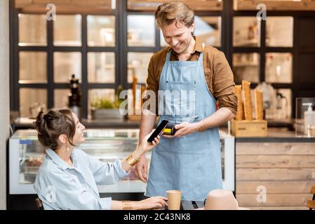 Glücklicher Kellner mit einem Kunden im Café oder Eisdiele, kontaktlose Bezahlung mit einem Smartphone Stockfoto