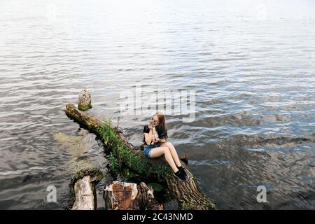 Junge blonde Frau mit blauen Haaren, Brille, Jeans-Shorts und einem schwarzen T-Shirt. Sitzt auf einem schwimmenden Baum in der Nähe des Sees mit einem drei-Monate-alt Stockfoto